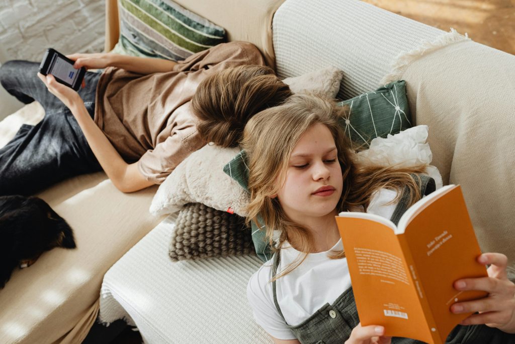 Two teenagers relaxing at home, reading and using a tablet on the couch.